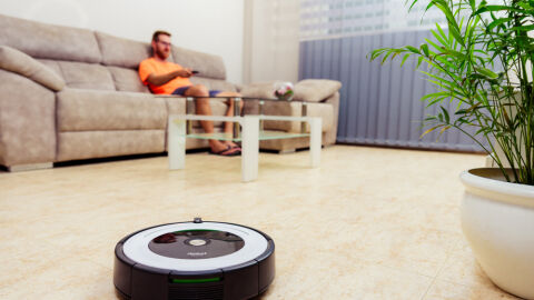 Un joven mira la televisión en el salón de su casa mientras un robot aspirador recorre el suelo