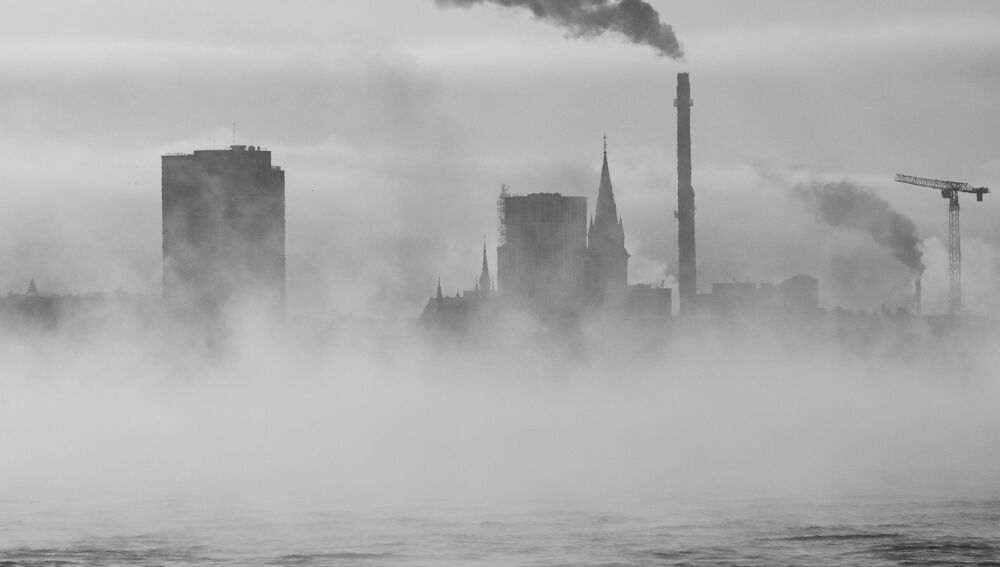 Paisaje urbano monocrom&aacute;tico con niebla y chimeneas industriales sobre el agua.