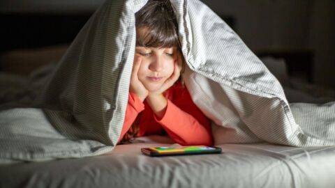 Los adolescentes que duermen menos de 8 horas son más proclives a la obesidad Niña bajo la sábana viendo un dispositivo móvil