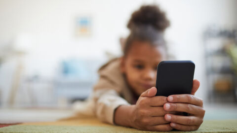 ¿Podemos usar los ‘chatbots’ como un amigo o un psicólogo? Cerca de una linda niña afroamericana sosteniendo un smartphone en el suelo en el acogedor interior de su casa, espacio para copiar