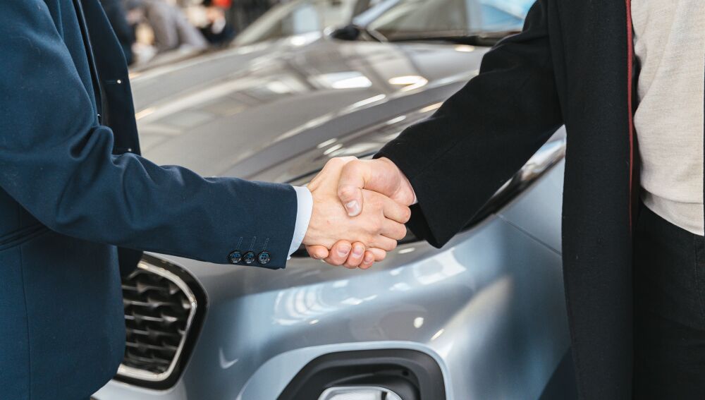 Un primer plano de dos personas estrechando manos frente a un coche en un ambiente interior Un primer plano de dos personas estrechando manos frente a un coche en un ambiente interior