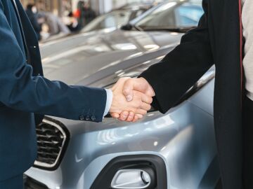 Un primer plano de dos personas estrechando manos frente a un coche en un ambiente interior Un primer plano de dos personas estrechando manos frente a un coche en un ambiente interior