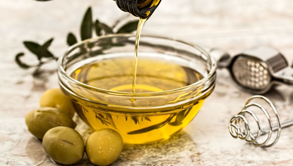 Primer plano de aceite de oliva siendo vertido en un tazón de cristal rodeado de aceitunas frescas y utensilios de cocina. Primer plano de aceite de oliva siendo vertido en un tazón de cristal rodeado de aceitunas frescas y utensilios de cocina.